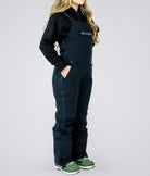 Woman wearing Nexarina Everette women’s black bib snow pants, side profile showing tailored fit and full-length leg coverage.