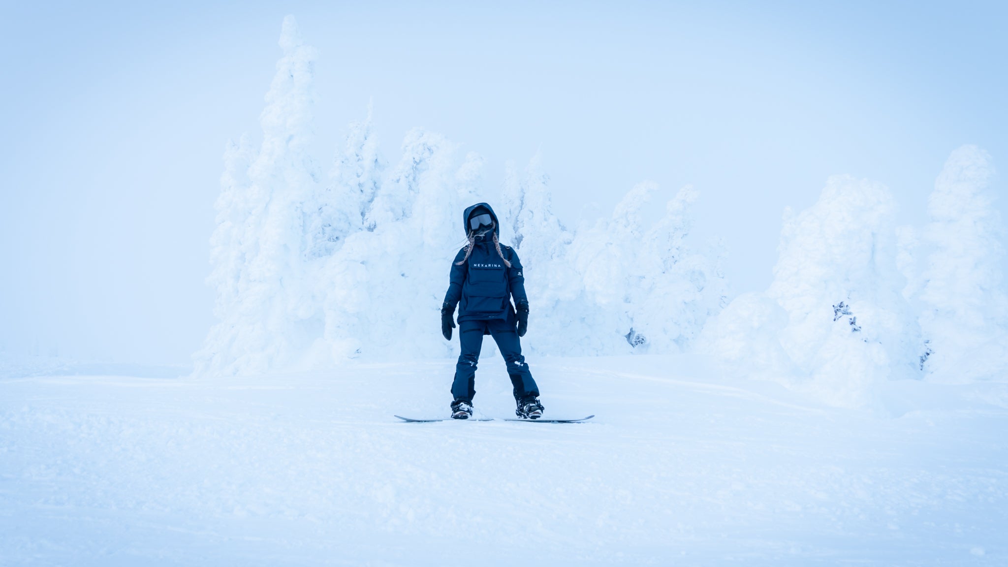 A snowboarder wearing a navy blue Nexarina jacket stands on a snowy slope, ready to ride. Nexarina outerwear is designed for all body types and performance.