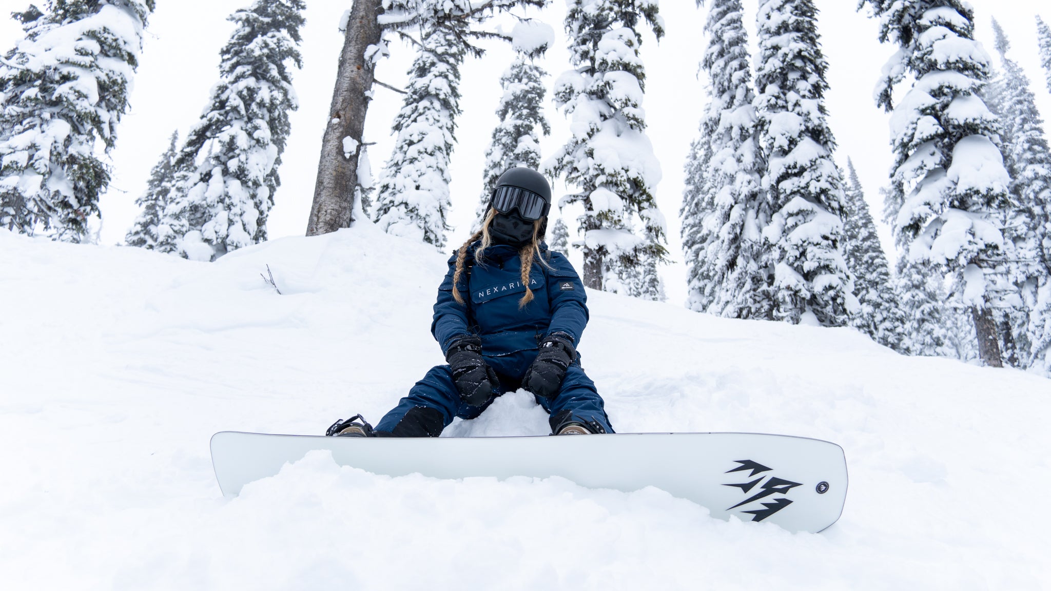 A snowboarder wearing a navy blue Nexarina jacket sits in the snow, surrounded by snowy trees. Nexarina outerwear is designed for all body types and performance.