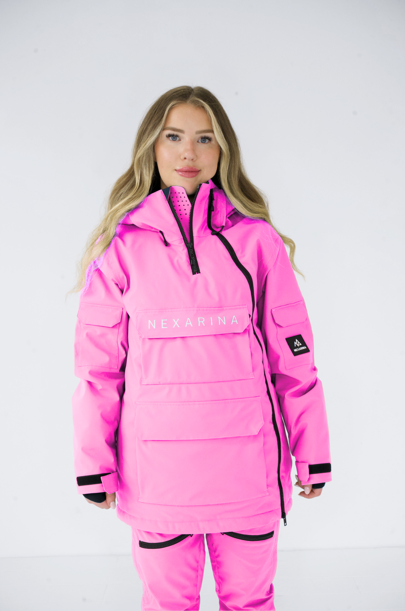 Woman smiling while adjusting hood of pink Nexarina jacket, front view, standing against a white background.