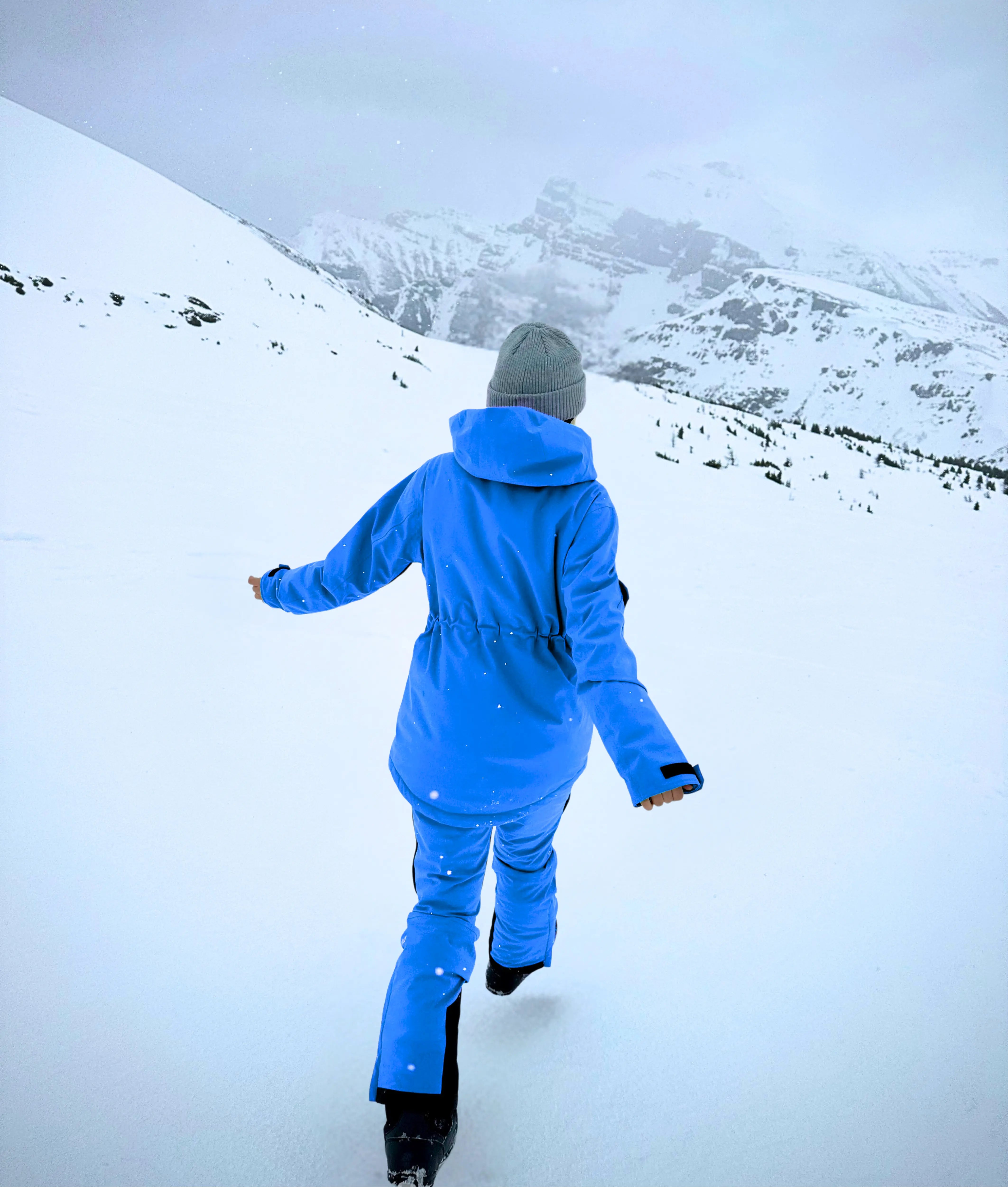 A model walks through the snow in blue Avani snow pants, featuring an adjustable waist, insulation, and flexible design.