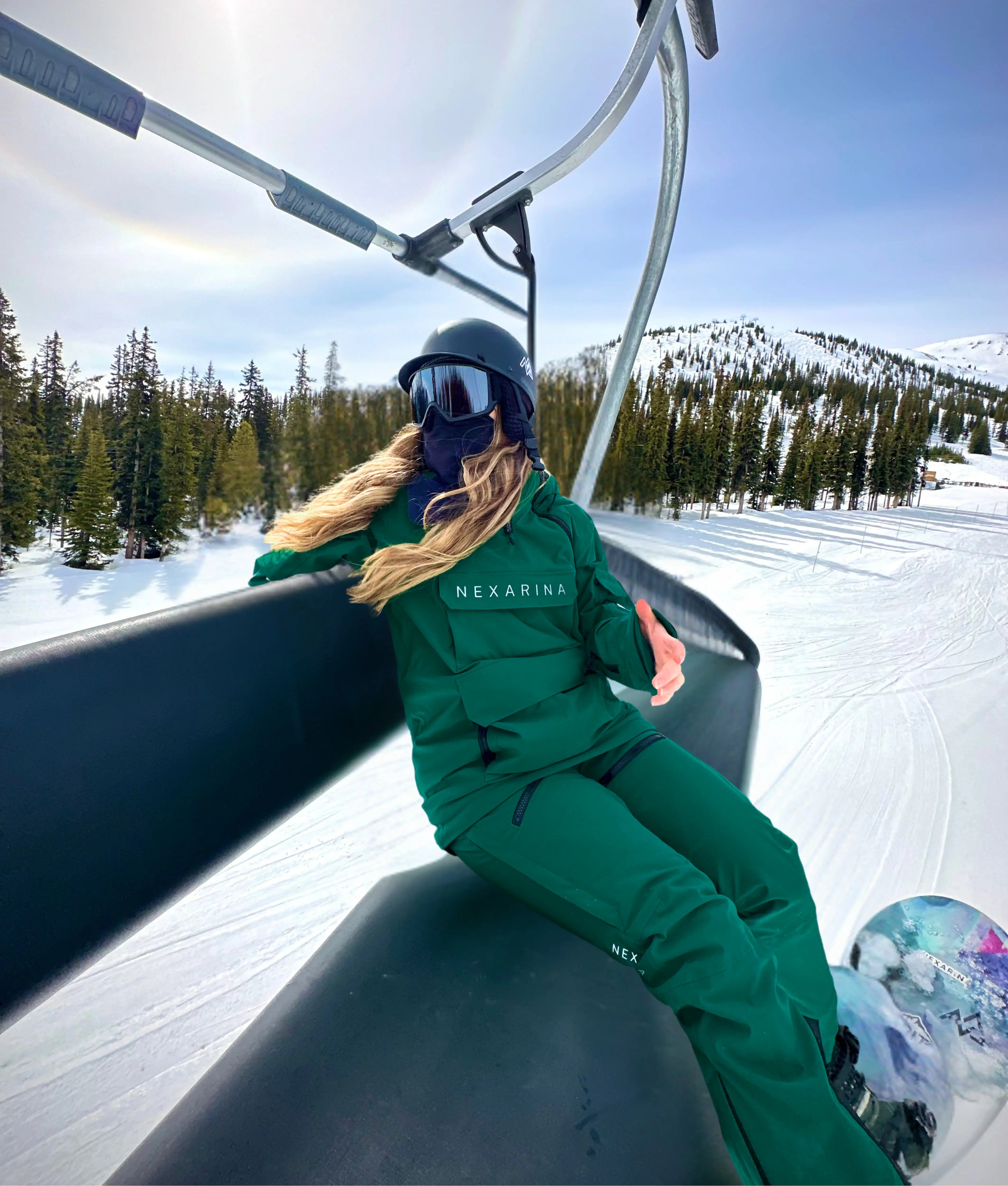 Female snowboarder in Nexarina Avani Ermerlad Green Snow Pant. She's sitting on a ski lift with snow-covered mountains and trees in the background.