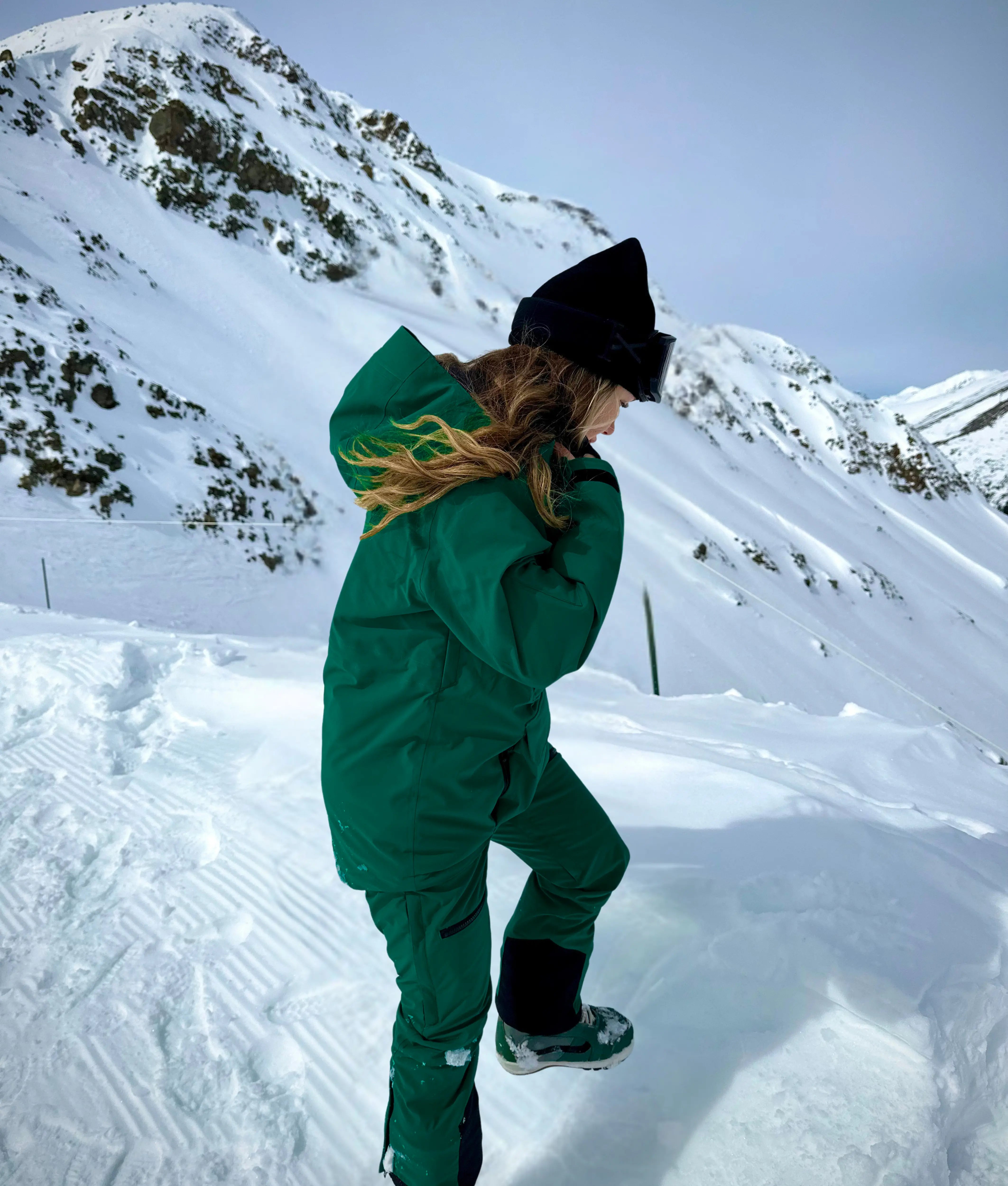 Woman wearing Nexarina Avani snowpants in emerald green standing in deep snow with mountain backdrop.