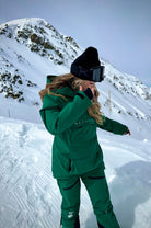 Woman wearing Nexarina hunter green Avani snowpants on snowy mountain, ski goggles on forehead.